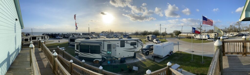 Panoramic view of Cedar Cove RV Resort with recreational vehicles parked in rows, an American flag and a Texas flag flying in the breeze, under a vast sky with the sun low on the horizon.