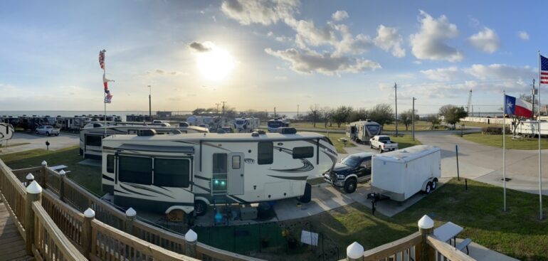 Panoramic view of Cedar Cove RV Resort with recreational vehicles parked in rows, an American flag and a Texas flag flying in the breeze, under a vast sky with the sun low on the horizon.